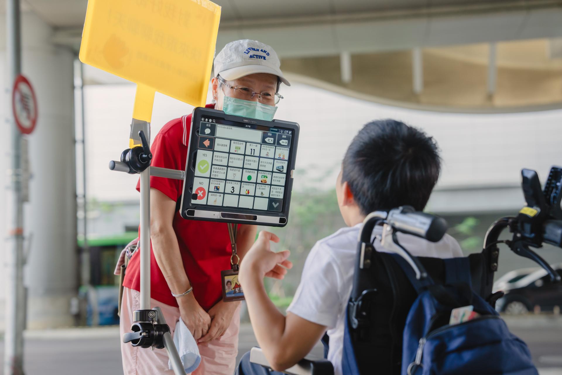 一位阿姨好奇地上前和朋寯聊天，朋寯用雙眼點選圖卡「我用眼動設備跟你聊天」回應，凱玫老師在一旁說明眼動的操作與溝通方式。